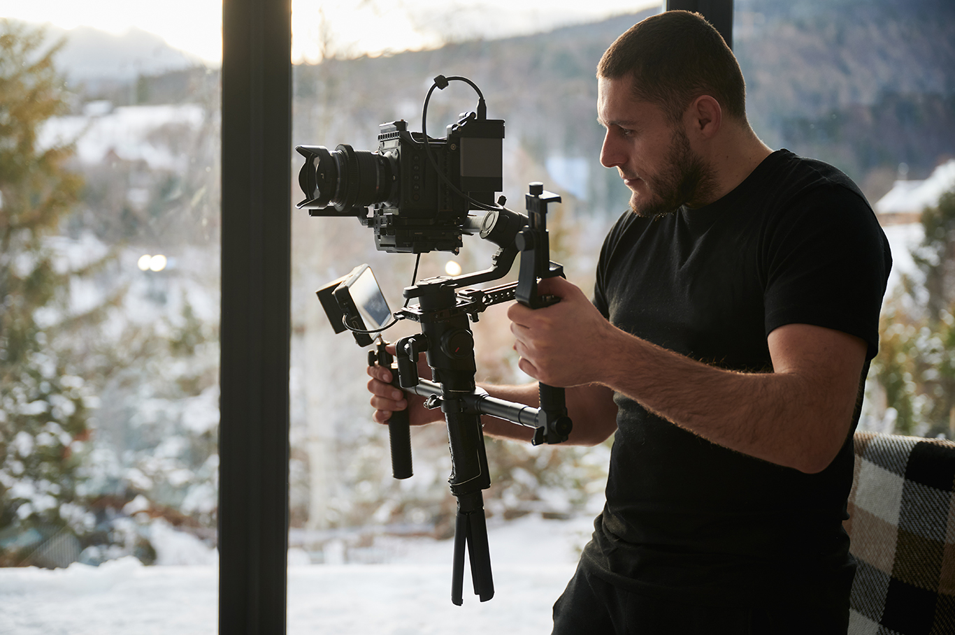 Videographer man shooting footage, using camera mounted on gimbal stabilizer equipment. Panoramic windows with winter landscape on background.